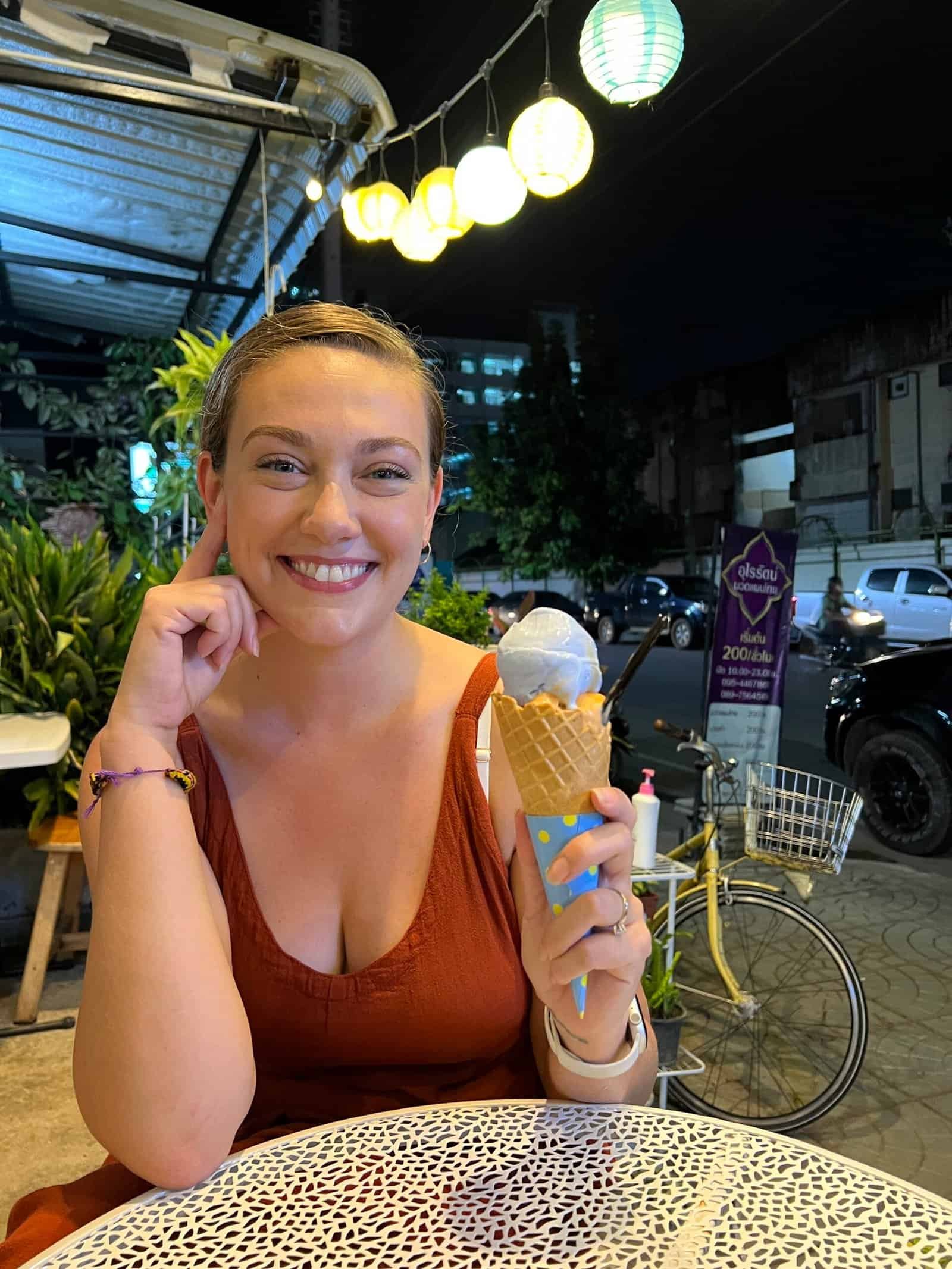 Ashley sitting at an outdoor café at night in Bangkok holding an ice cream cone, with street lights and traffic behind her