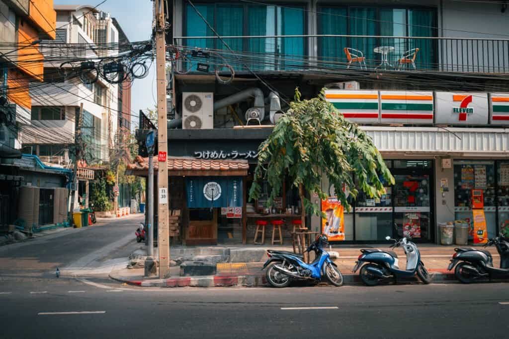 Bangkok side street with a 7-Eleven, scooters parked along the road, and overhead power lines
