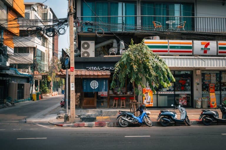 Bangkok side street with a 7-Eleven, scooters parked along the road, and overhead power lines
